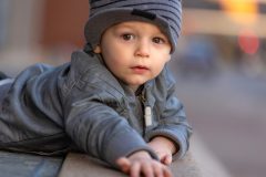 Toddler boy wearing a grey jacket and hat looking into the camera with bokeh in the background