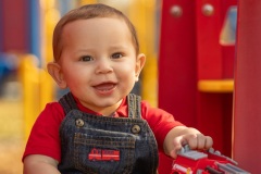 Infant boy in red shirt and denim overalls smiling and holding a toy firetruck