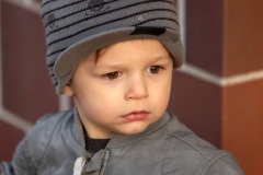 Toddler boy wearing a grey leather jacket stand against a brick wall